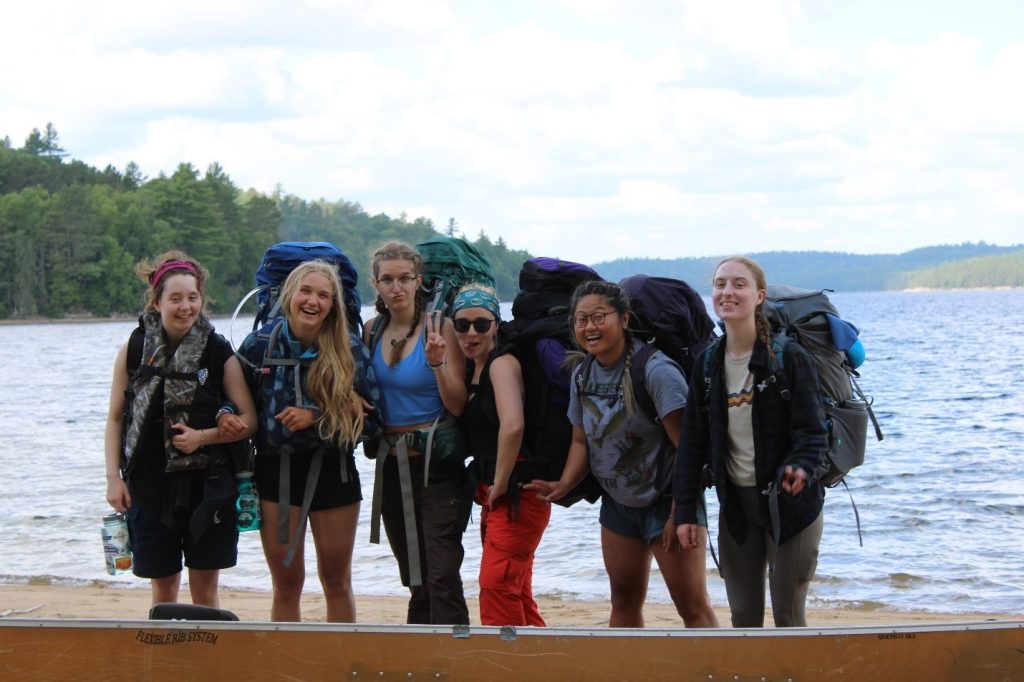 Six staff members off-hours, wearing backpacking gear, standing on a beach, smiling and goofing around