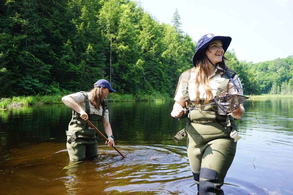 Two Discovery rangers dip-netting in a pond. The one on the right turns to the right and is caught mid-smile