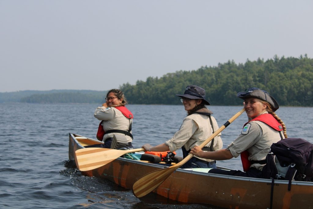 Three staff members paddling a canoe; they all turn their heads towards the camera and are smiling mid-paddle