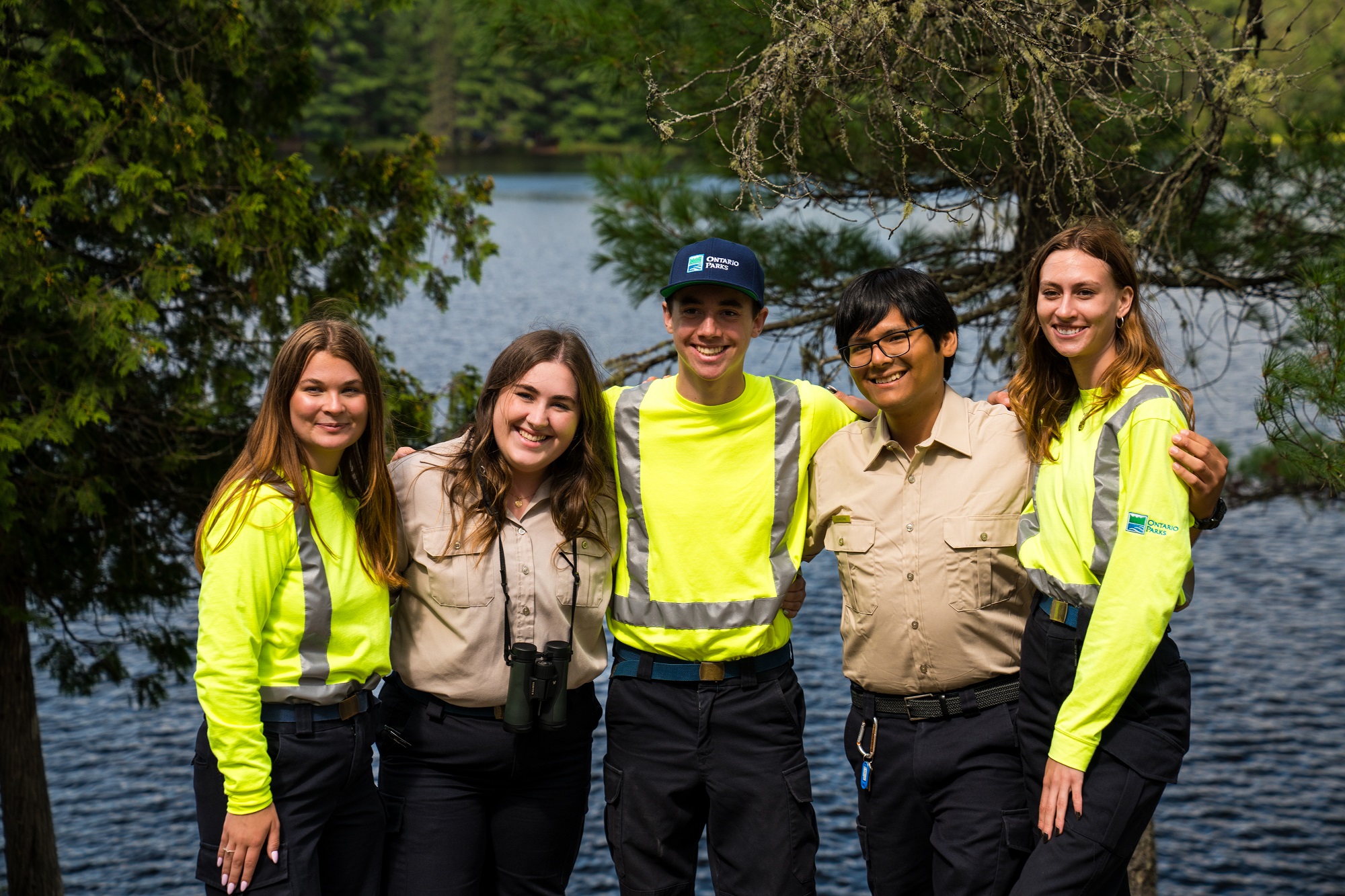 A group of smiling summer students, some wearing office uniforms, some wearing maintenance uniforms, standing on a beach in front of a river, their arms over each other's shoulders