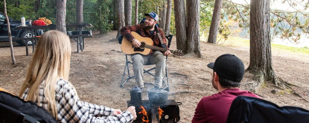 group sitting around campfire on campsite, one person playing guitar