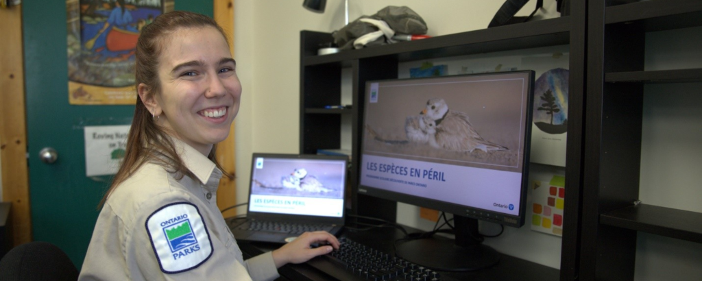 Ontario Parks staff member sitting at a desk in front of two computer screens on which a PowerPoint presentation slide from a grade four species at risk program is displayed.