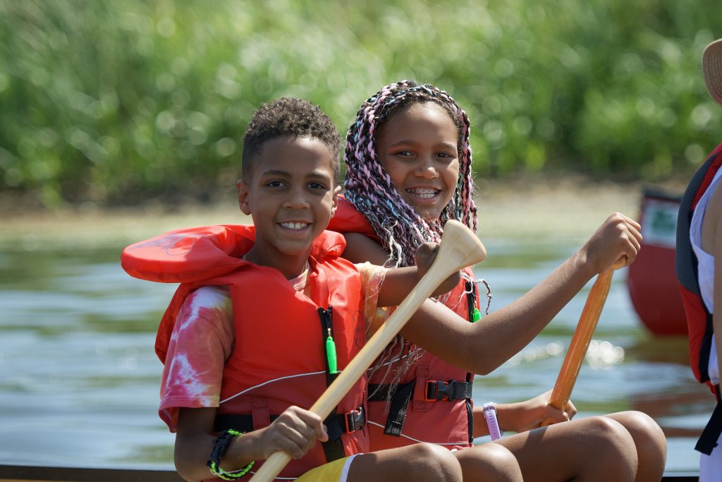two kids wearing lifejackets, holding paddles in canoe