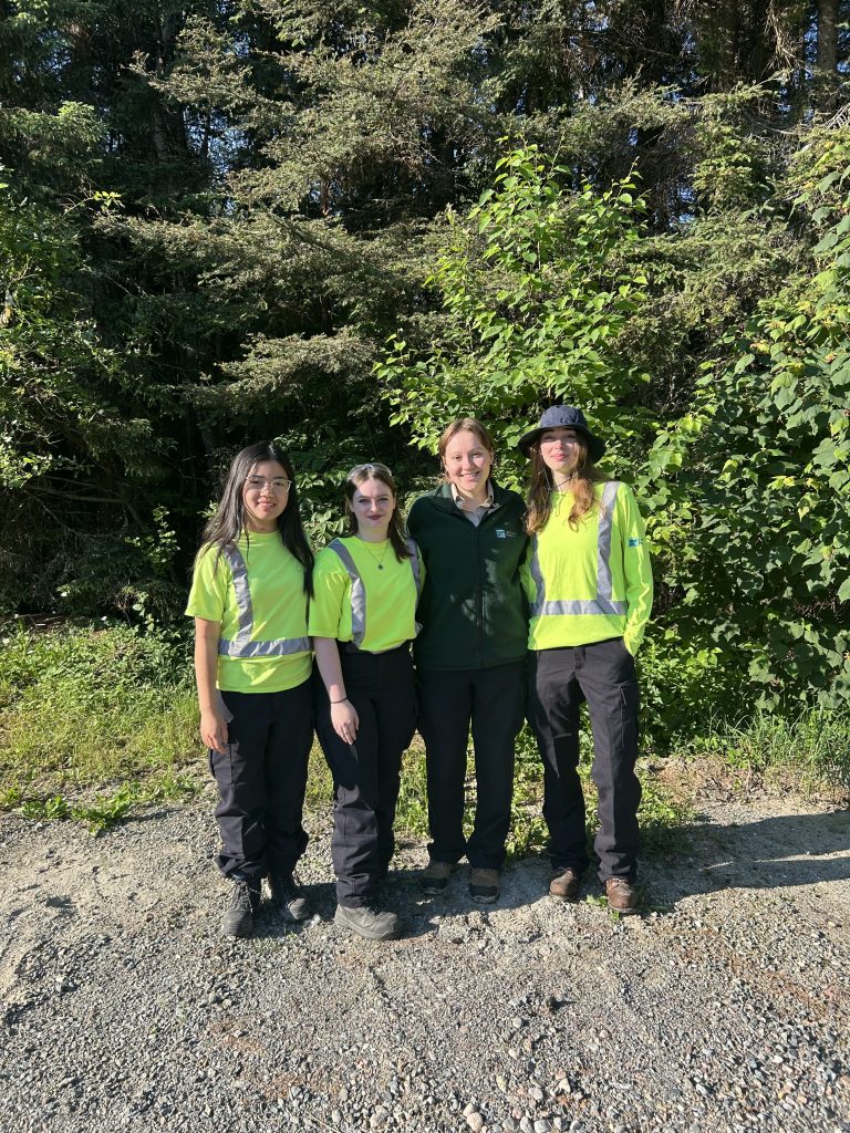 Emily, Ava D., Hannah, and Ava G. standing against treeline, three wearing high vis, one wearing fleece sweater