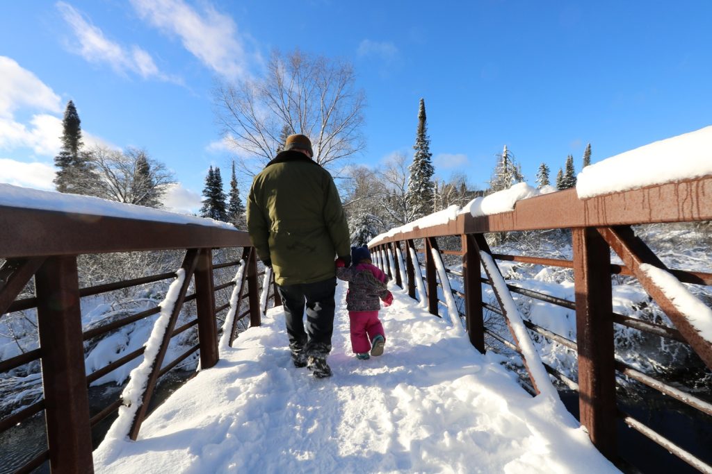 parent and child walking over a snowy bridge toward the forest