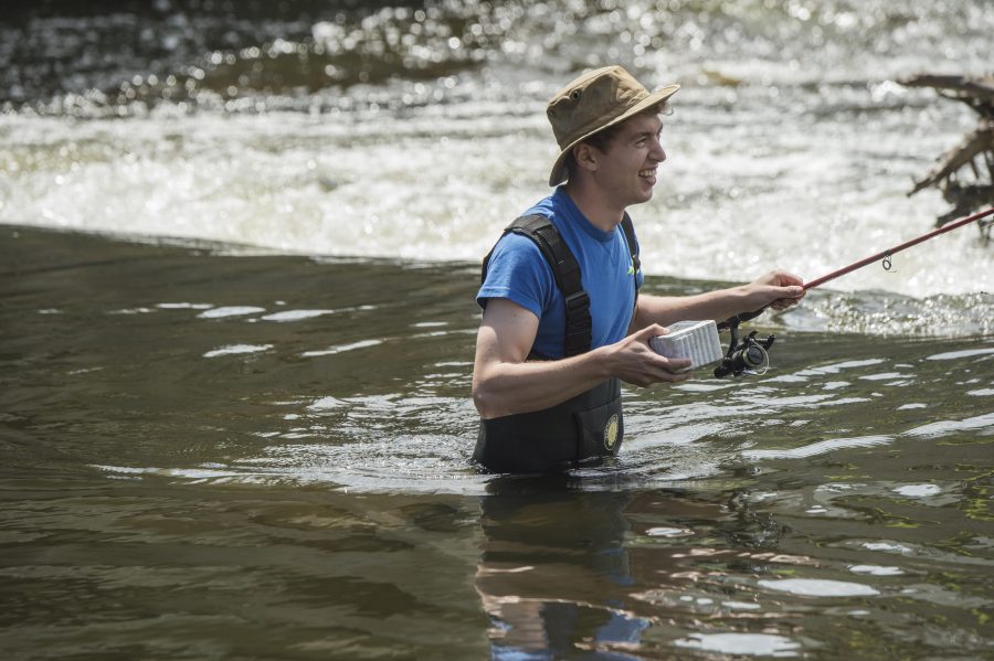 Man wades through the river holding worms and a fishing pole. 