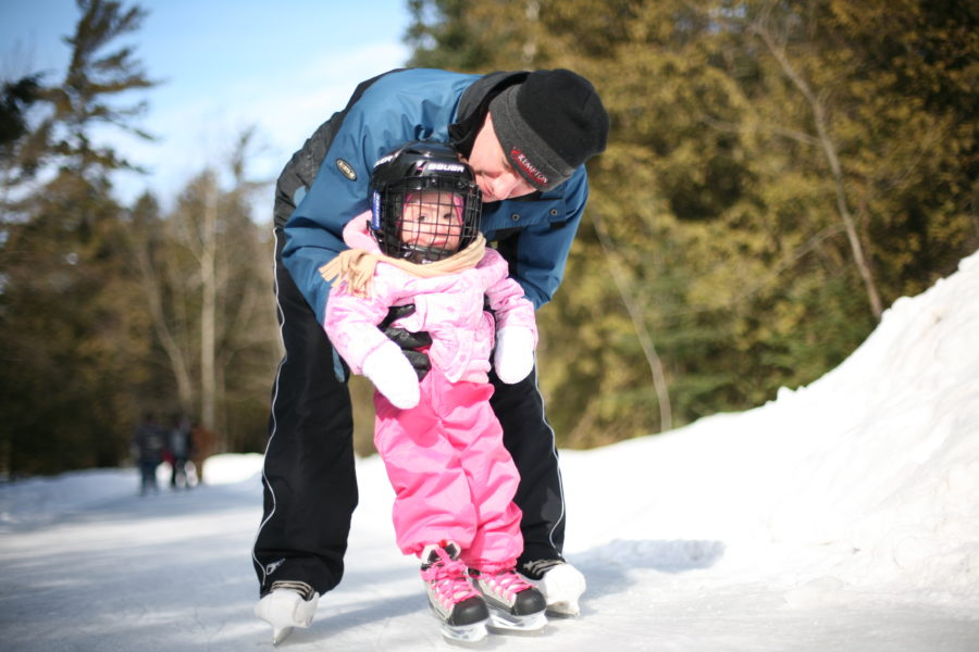 Dad with 2 year old daughter in pink snowsuit on skates on the skate trail