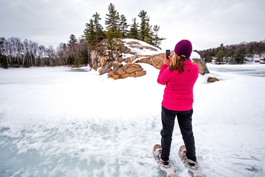 A person on snowshoes taking a photo of a frozen lake and snow-covered landscape with a pink rocky outcropping in the background