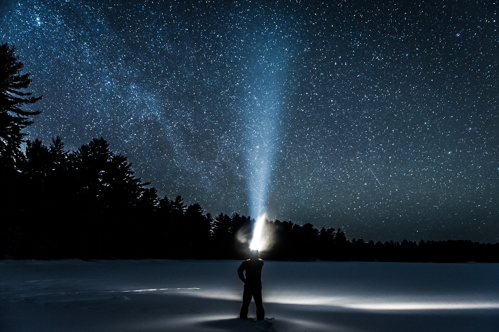 A person standing on snow shining a flashlight up into the starry night sky.