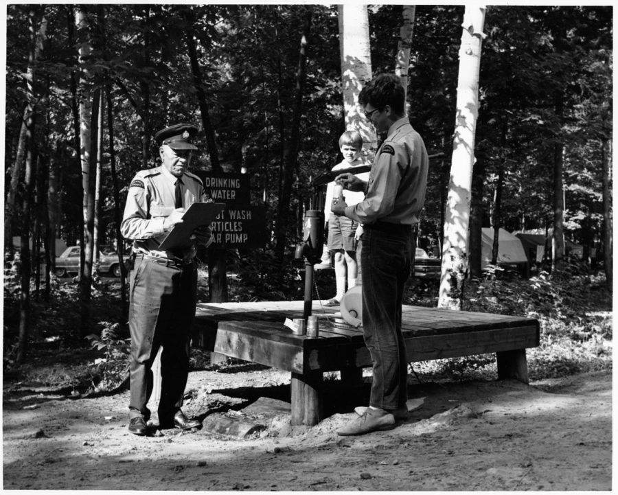 Older gent records something on a clipboard while younger guy samples water at a water station while a kid looks on in the background