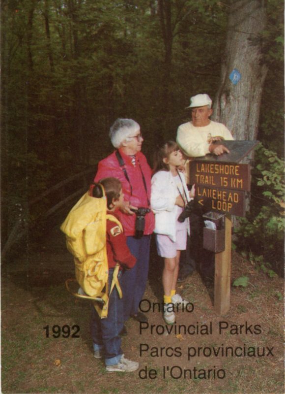 Photo of two older adults with two kids looking (all dressed in 90's fashions) at a trail map that is over a sign saying "Lakeshore Trail 15 km, Lakehead loop" . Text states "1992 Ontario Provincial Parks, Parcs provinciaux de l'Ontario".