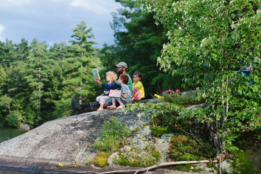 Guy with cap reading to three kids on a rock, facing the water