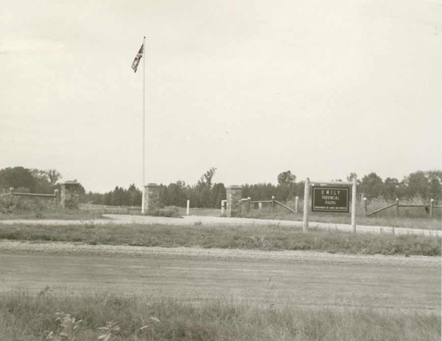 Black and white photo of a field with bramble, some fencing, a flag and a sign reading Emily Provincial Park