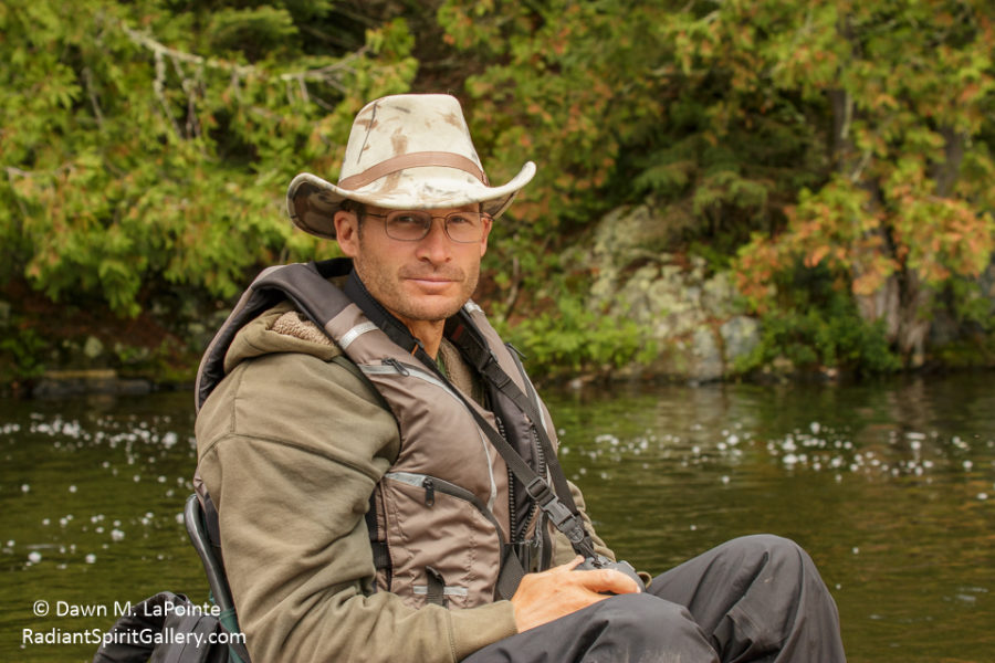 Guy sitting in a camp chair with a beige australian outback hat, life jacket, brown hoodies and grey quick dry pants with water and trees in the background