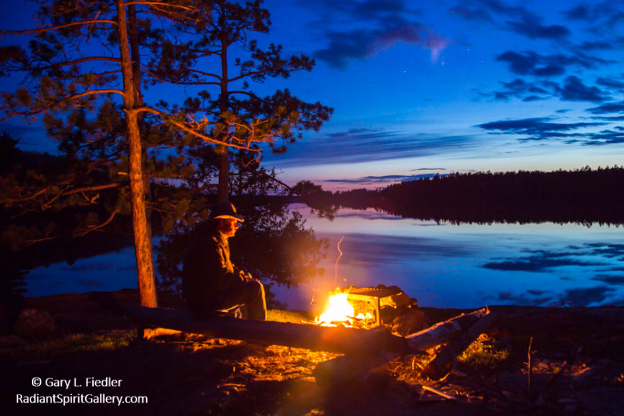 Guy with hat at dusk sitting in front of a fire with a sun set in the background