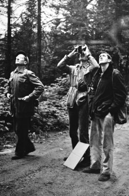 Three men in black and white photo looking up (one with binoculars)