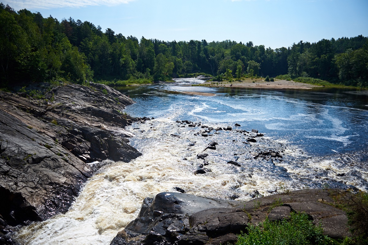 Narrow waterfall headed out of lake with sand beach and coniferous shore