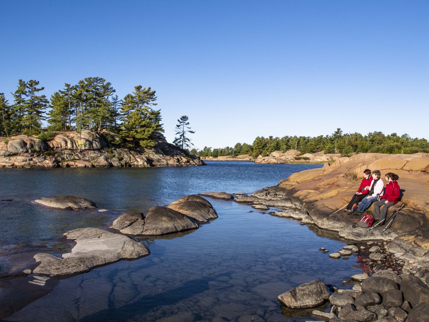 Three people sitting in the mid-distance on a rock shore of a calm, shallow body of water.