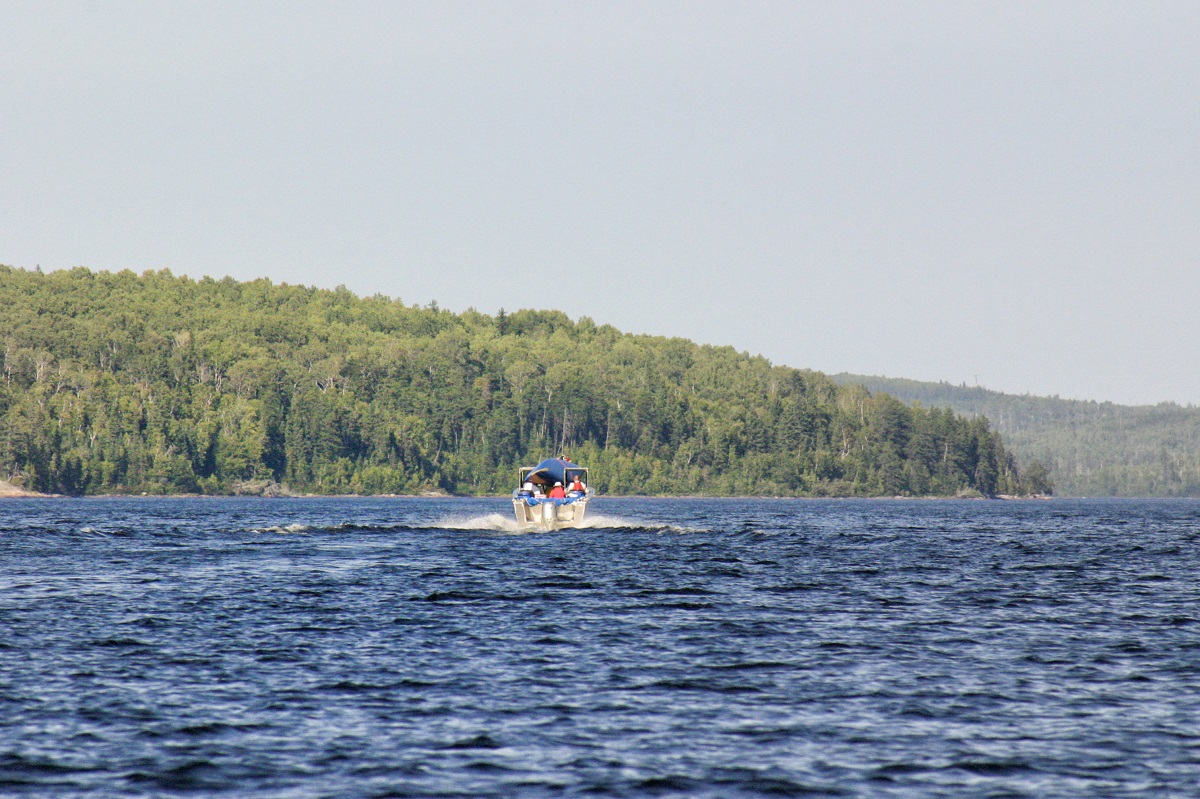 Blue lake with boat going away from photographer, forest and grey sky in the backgorund