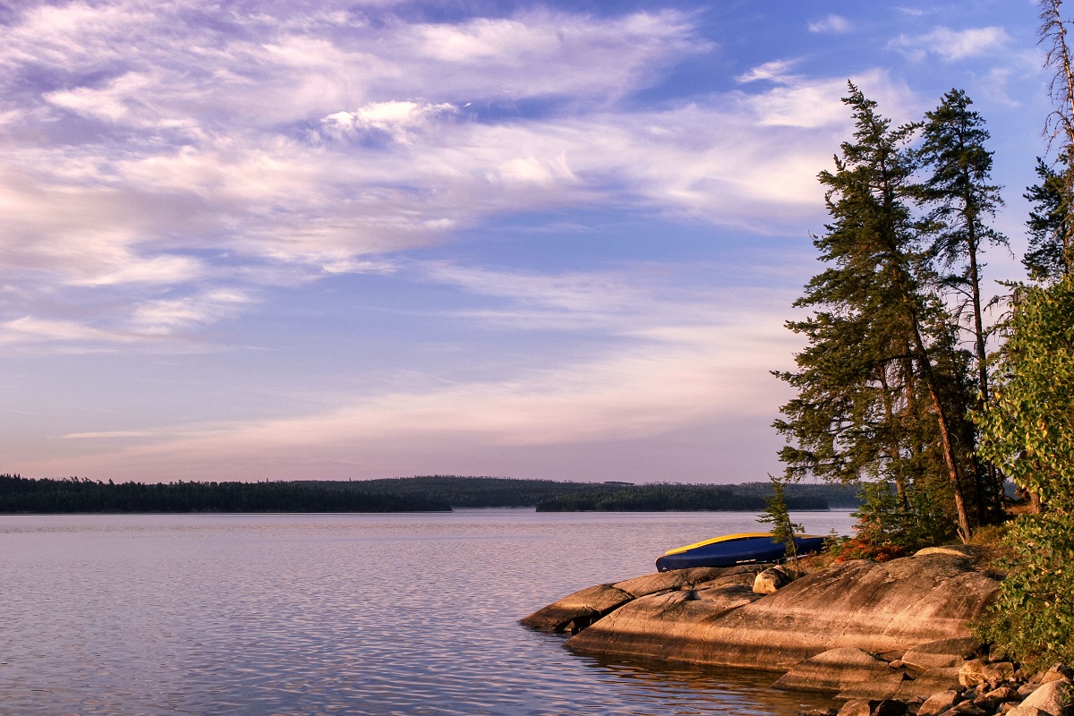 Rock shore with conifers on the side of large lake with blue whispy sky overhead