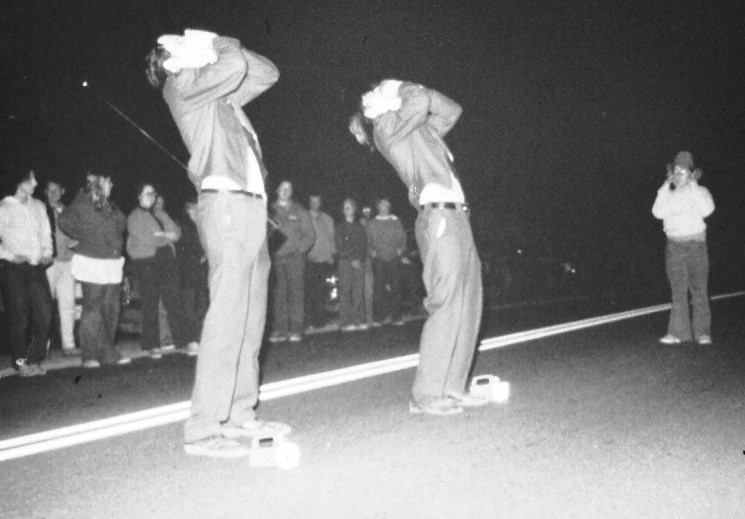 Two men howling up at the sky at night with people watching from behind