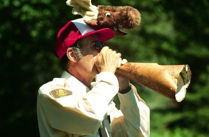 Guy with a red ball cap with a stuffed creature on top using a piece of rolled up birch bark as a megaphone