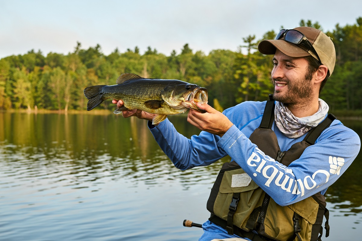 Guy with a cap and pfd, holding up and looking at a largemouth bass