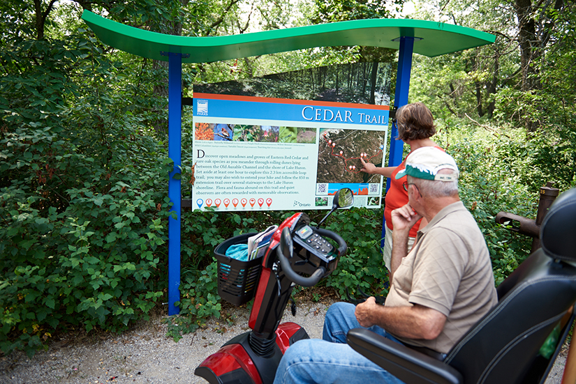 Man in scooter and woman stand in front of trail sign