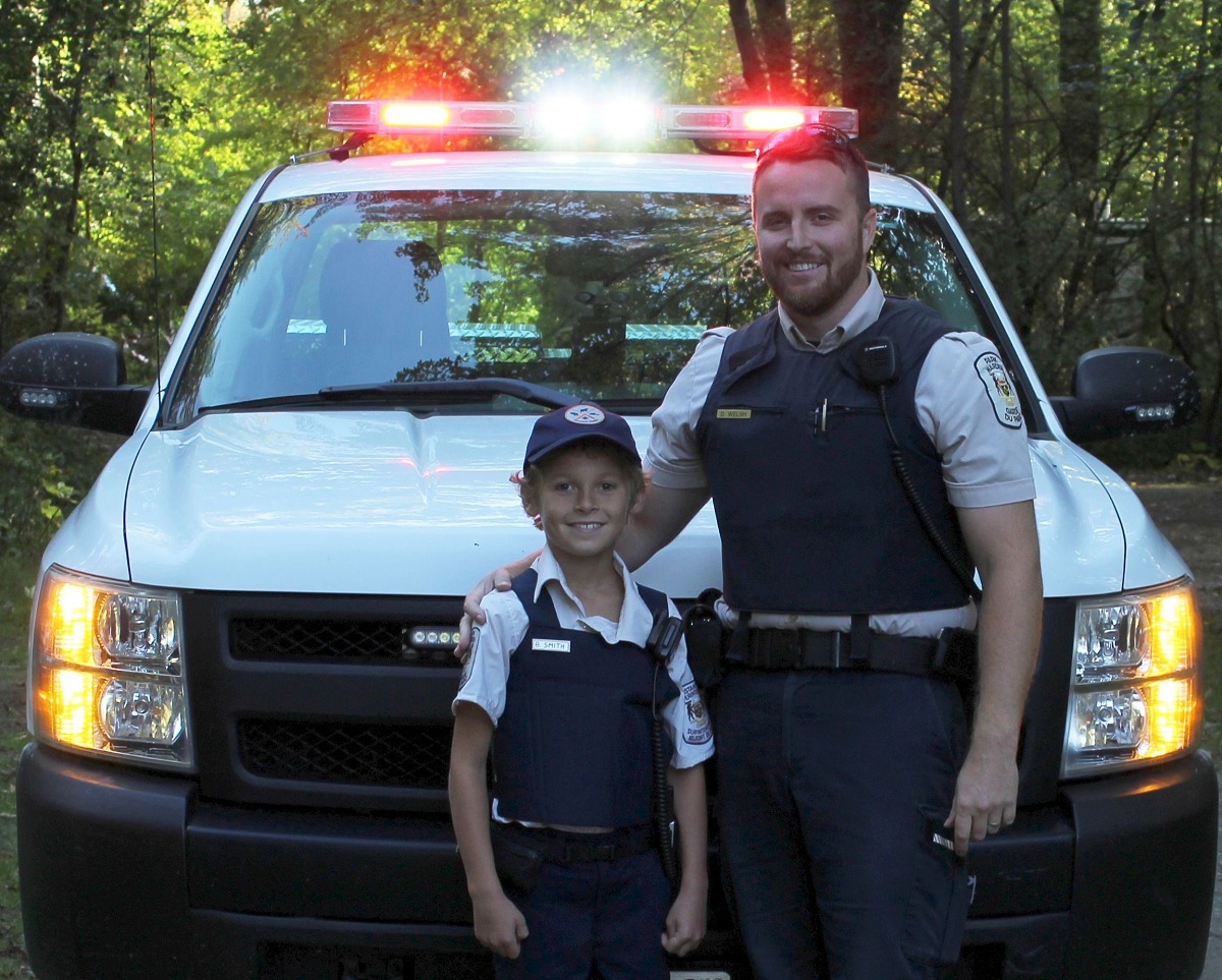 Park warden with child dressed as a park warden in in front of park vehicle with lights on the vehicle