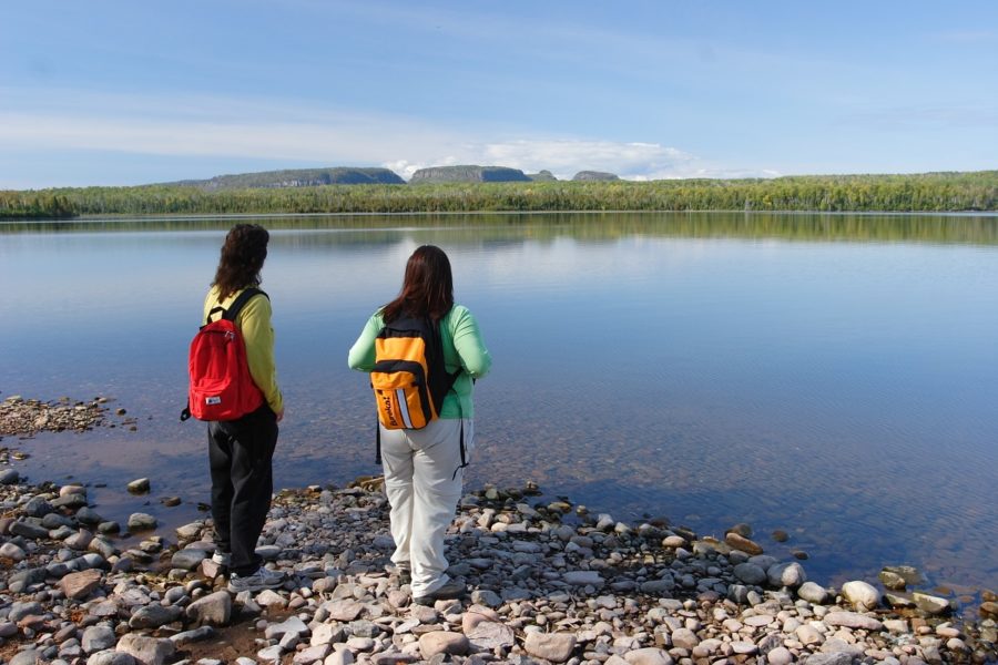 Two people with backpacks standing on a shore looking out at landform across water