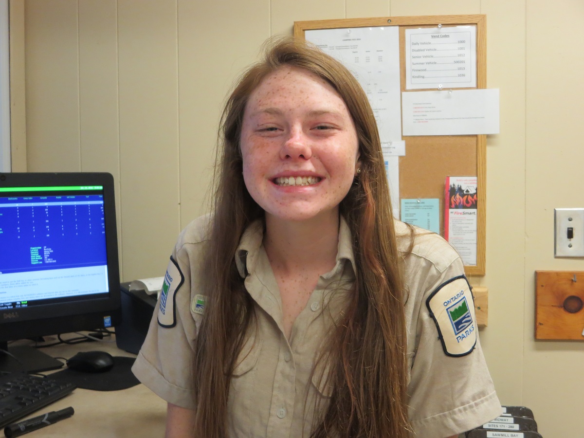 Girl with long hair in park uniform, sitting in front of a desk, smiling at the camera