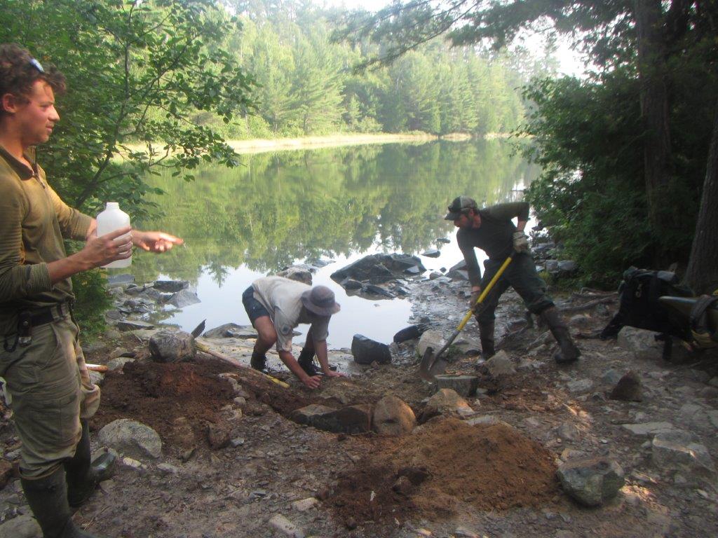 two men digging rocks and dirt while another drinks water