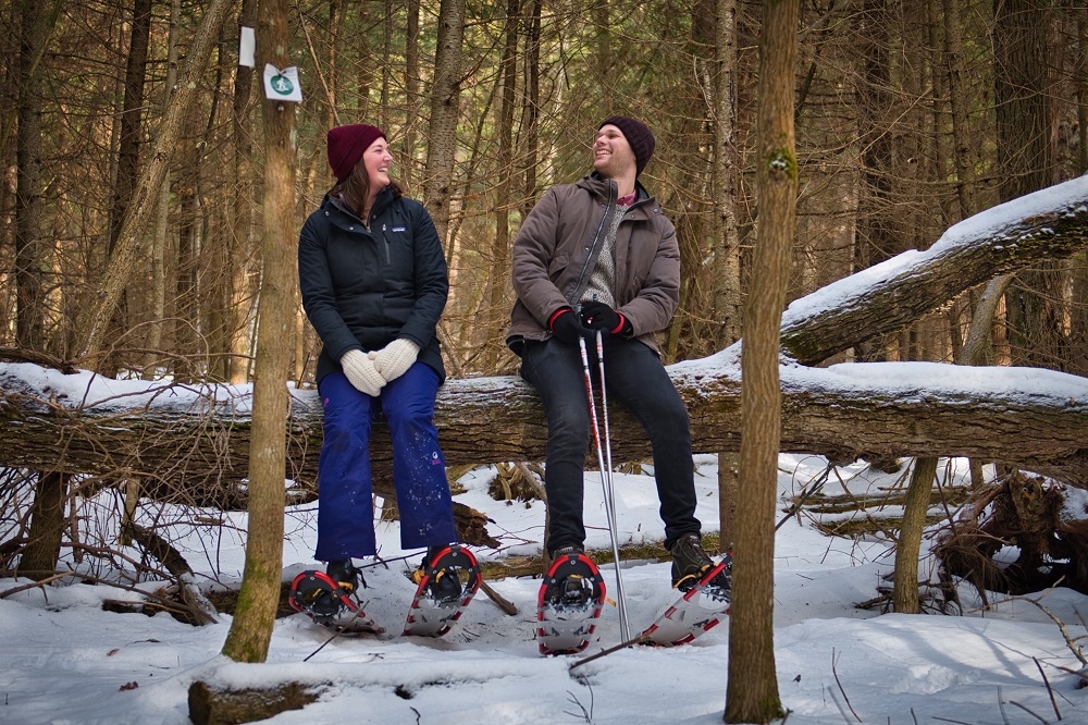 Two people sitting on a log in a snowy forest, wearing snowshoes and laughing