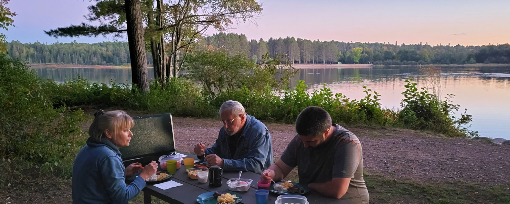 Three people having dinner at a picnic table at sunset on shore of a body of water