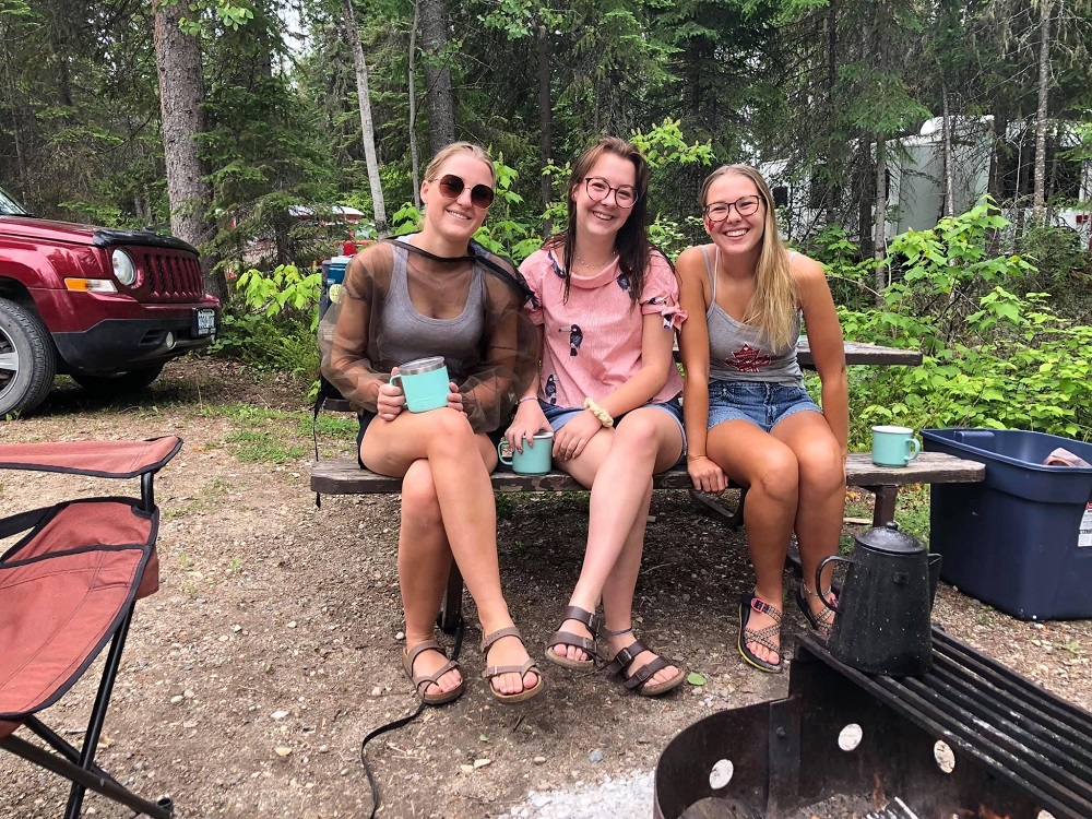 The three sisters sitting on the same picnic table on a campsite, smiling