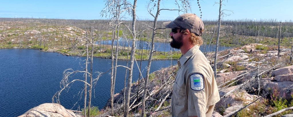 Staff standing on a rocky mountain in the middle of a burn landscape, a lake in the background