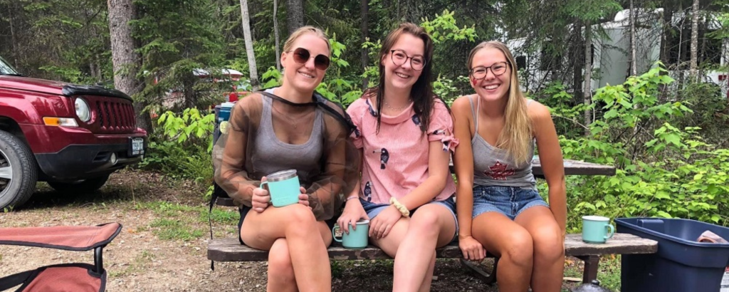 The three sisters sitting on the same picnic table on a campsite, smiling