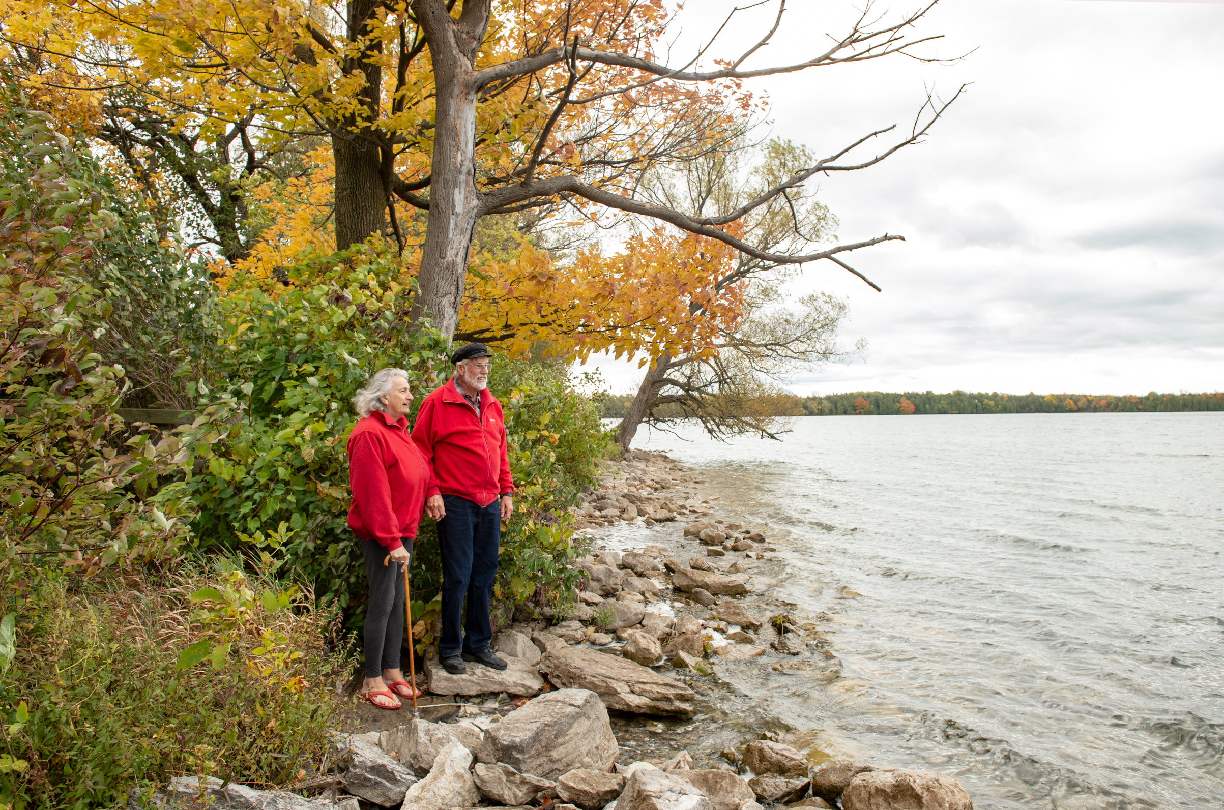 Two seniors wearing red jackets standing on the rocky shoreline of a lake in the fall