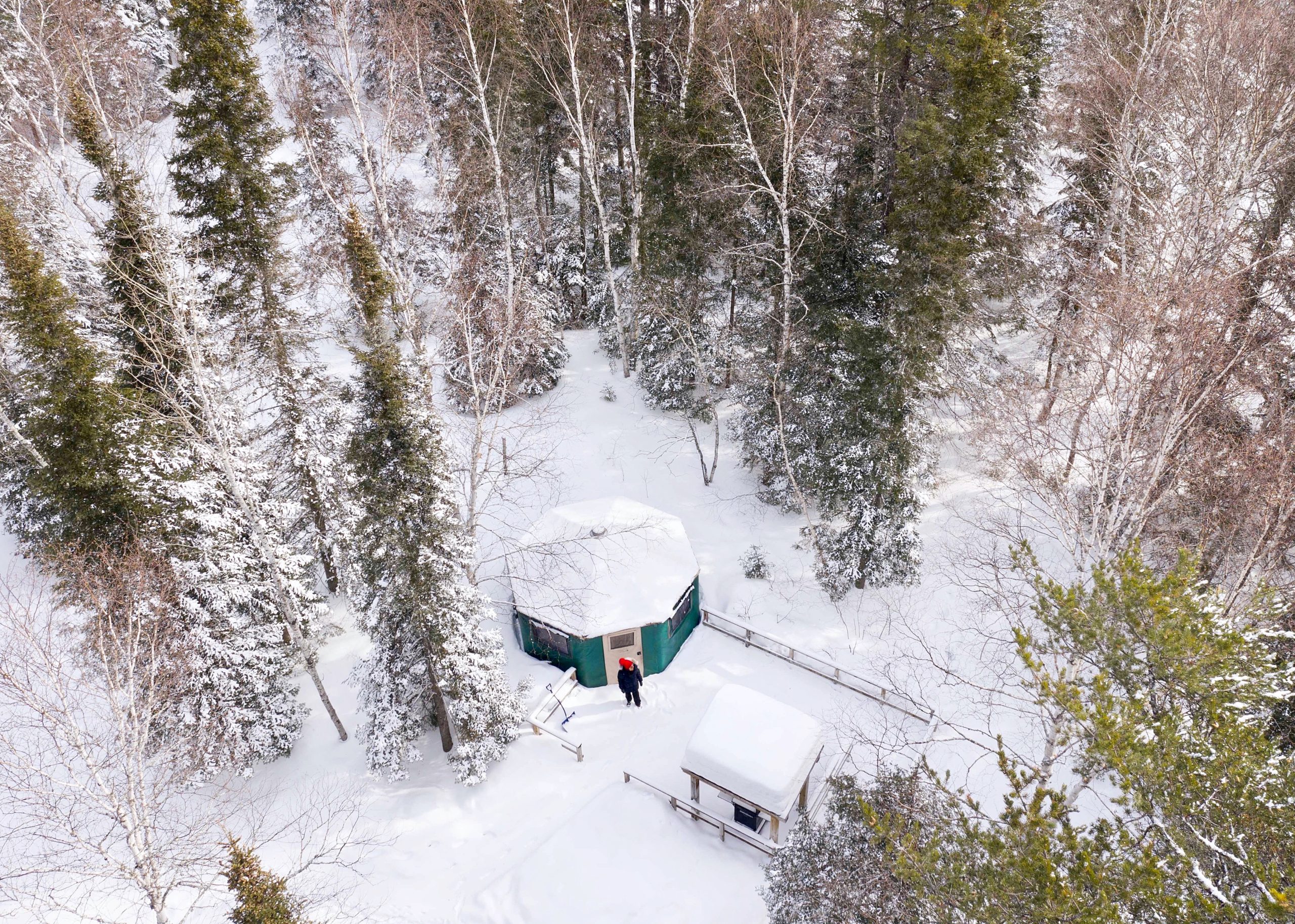 An aerial view of a snow-covered yurt in a forest, with a person standing in front of the door