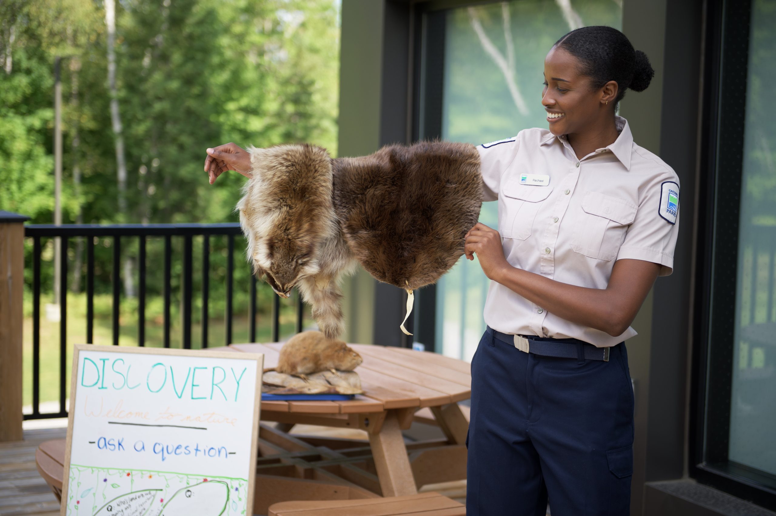 A Discovery ranger displaying an animal pelt on their arm in front of a building with a whiteboard beside them