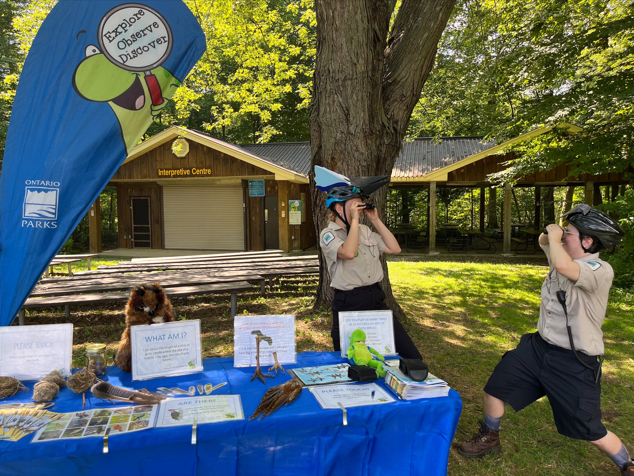 Two staff members wearing homemade props on their heads to look like birds staring at each other with binoculars in front of a bird-themed Discovery exploration station