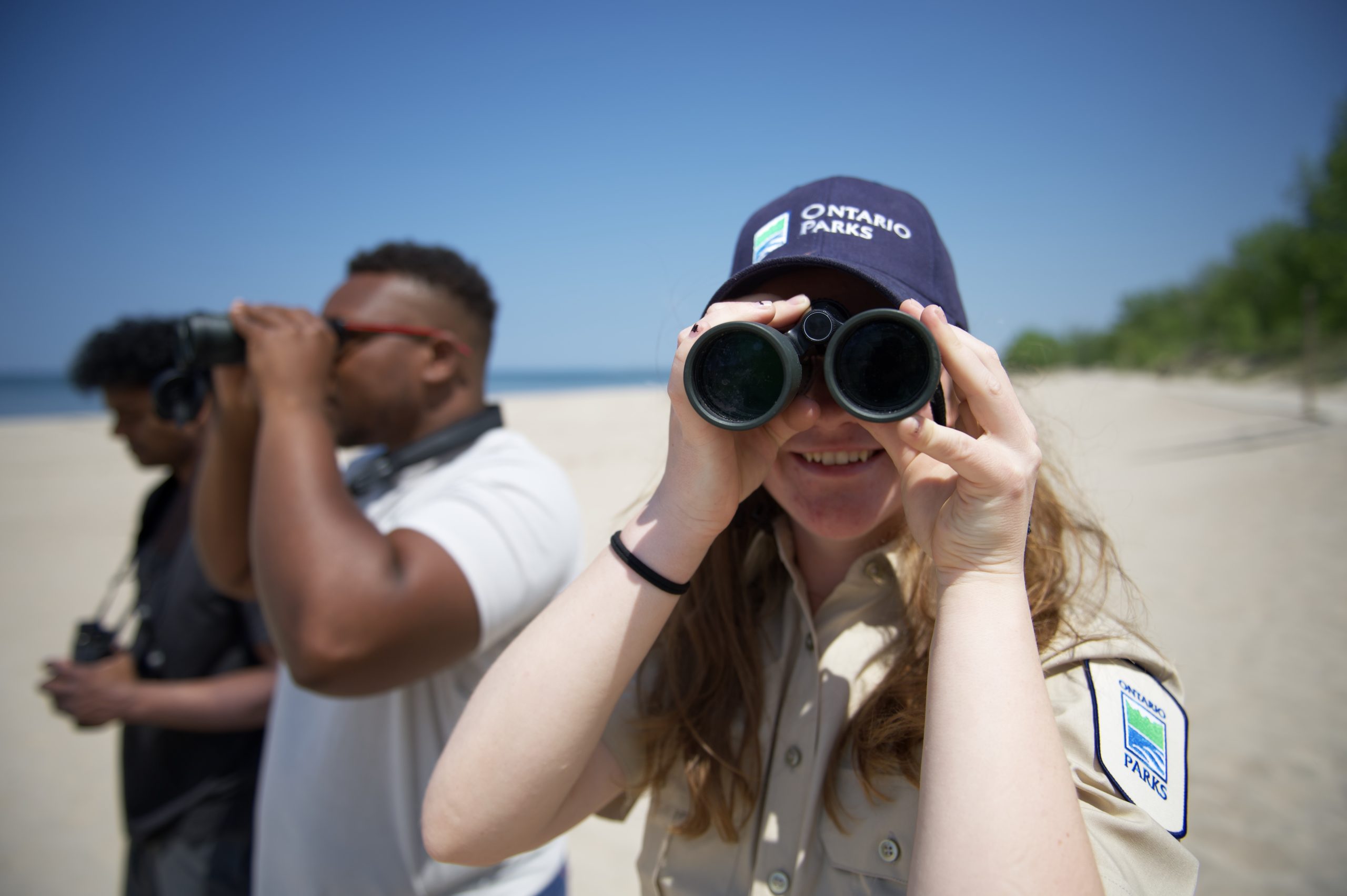 A Discovery ranger looking through binoculars on a beach beside park visitors who are doing the same