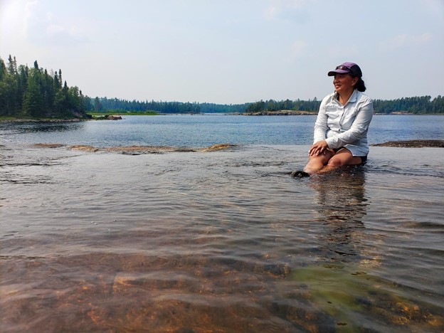 A person sitting in a shallow pool of water in a larger body of water, hemmed in by a forest in the distance.