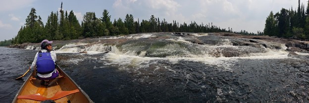 A canoe with a person in the front seat paddling towards a small, wide waterfall surrounded by trees