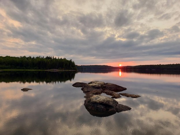 A red sun setting over the horizon, reflecting off a clear lake with large rocks breaking through the surface. The sky is cloudy and the clouds also reflect off the surface.