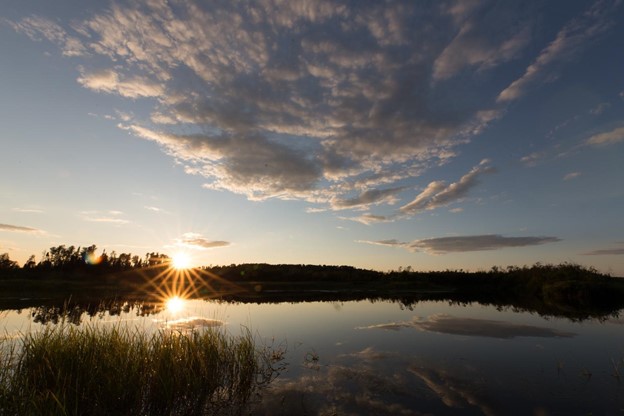 A yellow sun sitting low on the darkened horizon, reflecting off a clear lake. Above, the sky is dotted with white-grey clouds.