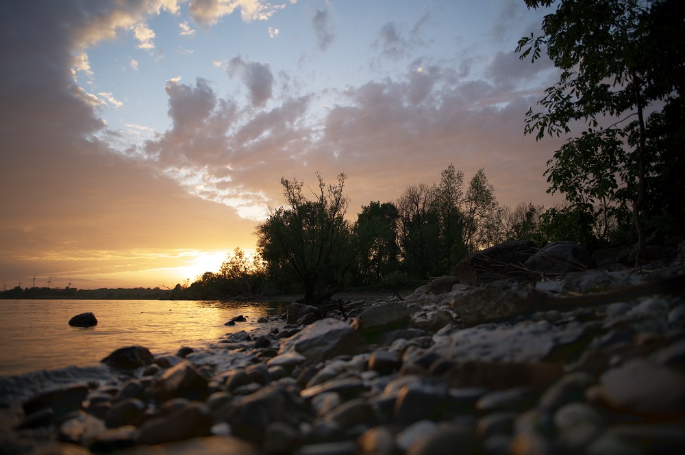 Low perspective image from rocky beach looking out over water at sunset. The sky is soft orange, purple and blue, with colourful clouds across the sky. 