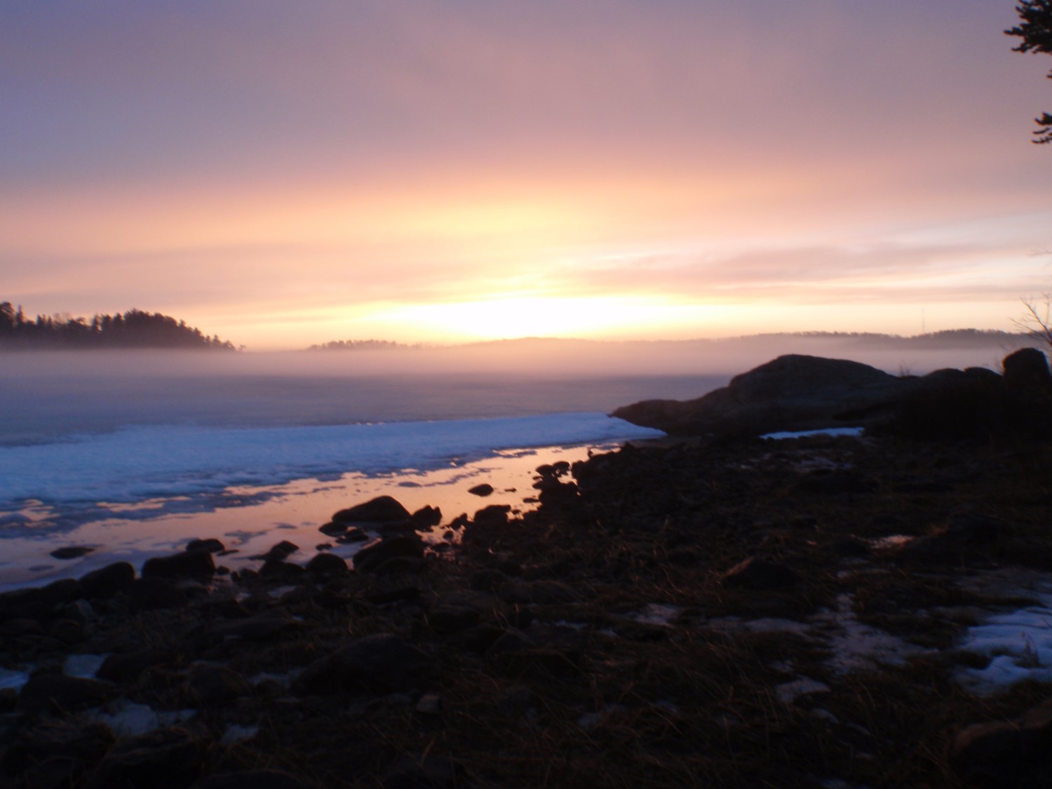 The sun setting in a light pink and purple sky behind a blue lake and darkened beach.