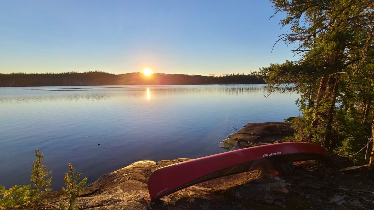 The sunsetting across from a lake. A red canoe is resting in the foreground.