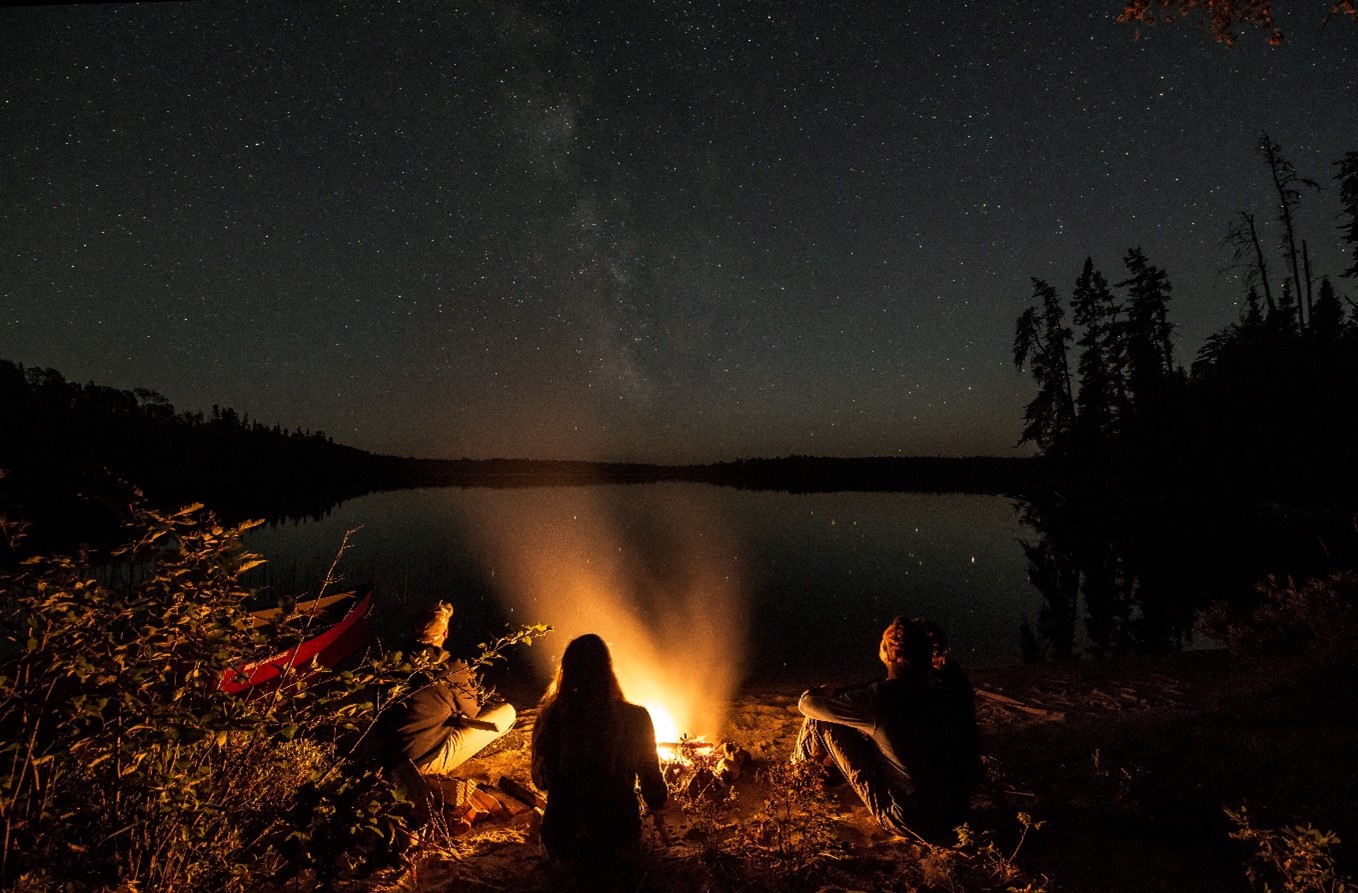 People sitting around a campfire, next to a lake, under a star-filled sky